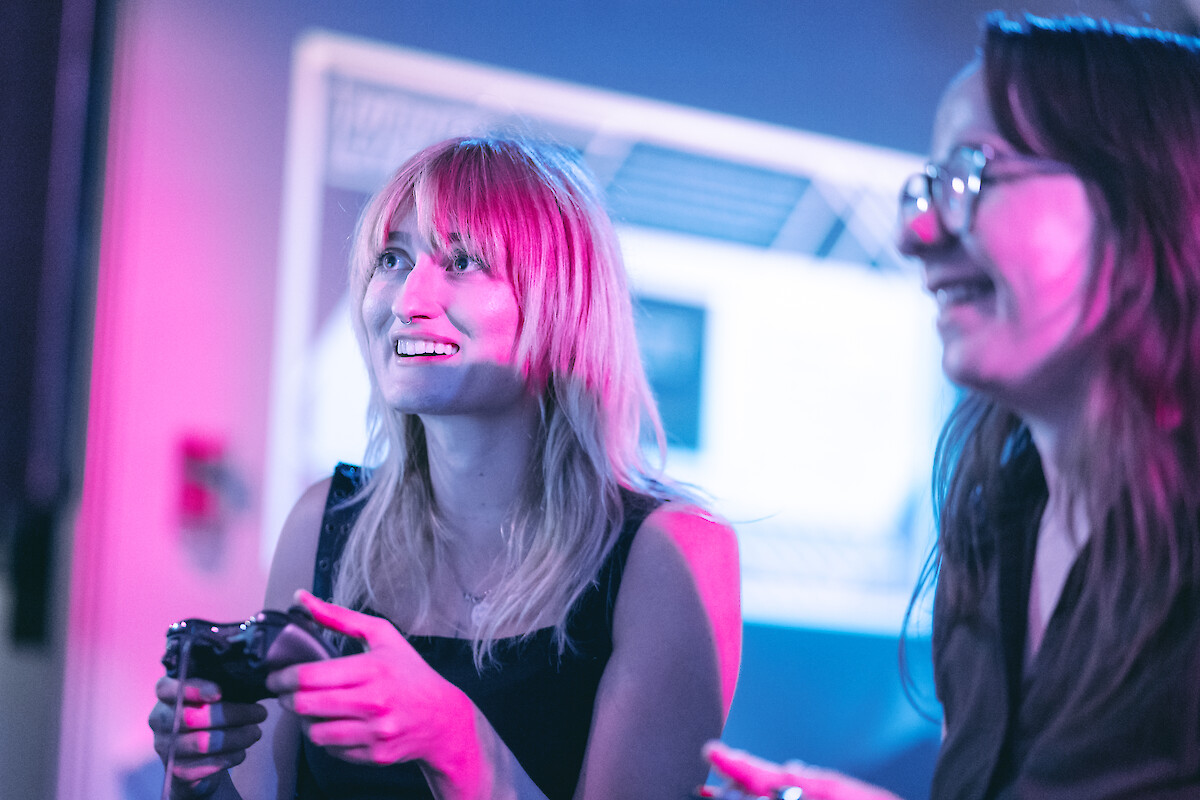 Two people are smiling and playing a video game in a room with a blue and pink light ambiance.