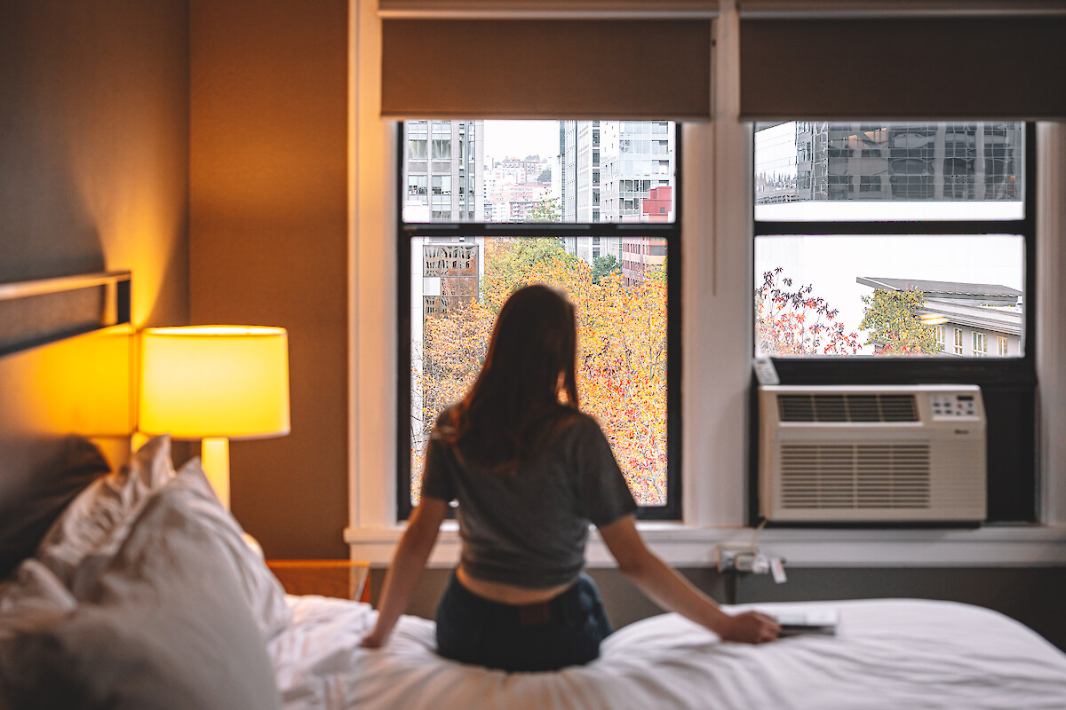 A person sits on a bed, looking out a window with urban and autumn scenery; a cozy room with a lamp and air conditioner, concludes the scene.