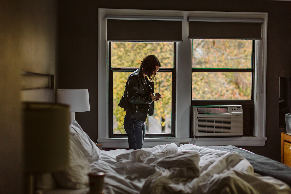 A person stands in a dimly lit room next to a bed, looking at a phone, with a window view of trees outside.