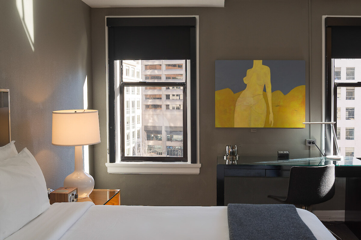A hotel room with a bed, a lamp on a bedside table, a window, a desk with a chair, and a painting on the wall.