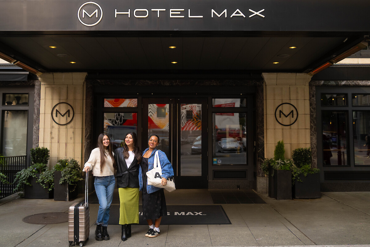 Three people stand in front of Hotel Max with one holding a suitcase, posing happily for a photo.