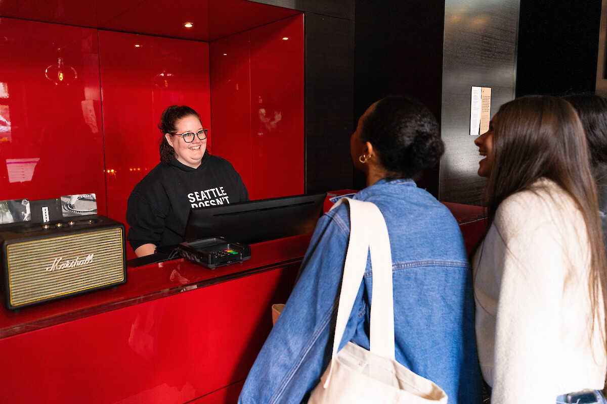 A person at a red counter smiling at three visitors; a Marshall speaker and card machine are on the counter.