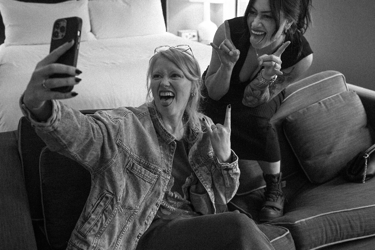 Two people seated on a couch in a cozy room taking a selfie and smiling, with a bed and a lamp in the background.