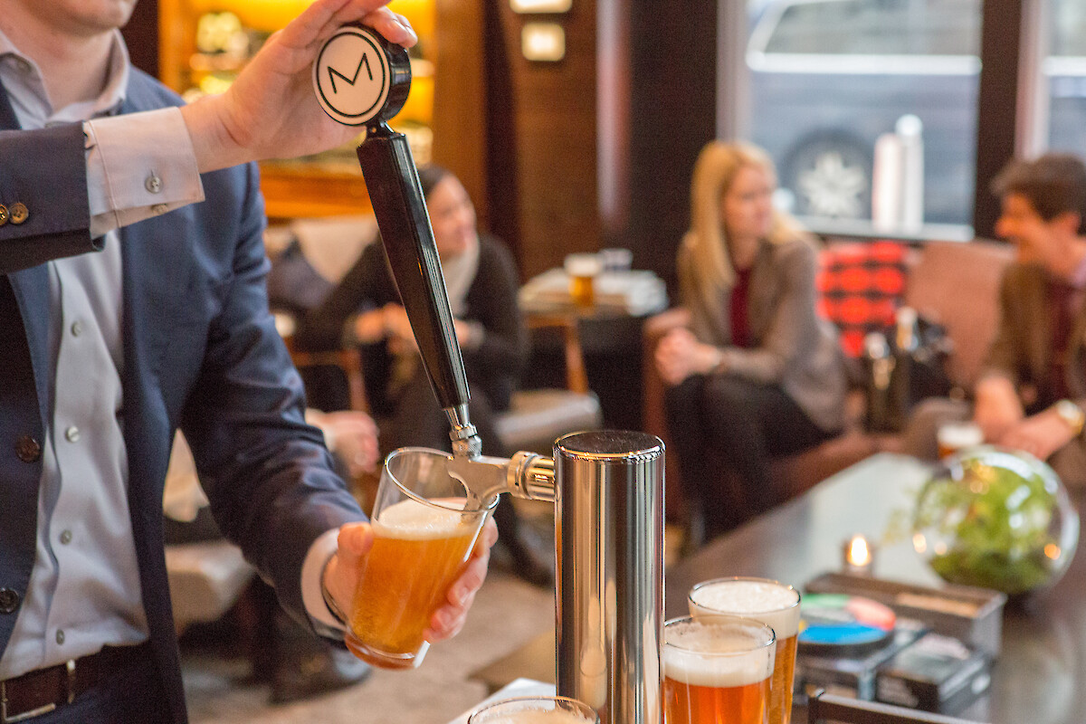 A person pours beer from a tap into a glass at a gathering, with people sitting and chatting in the background.