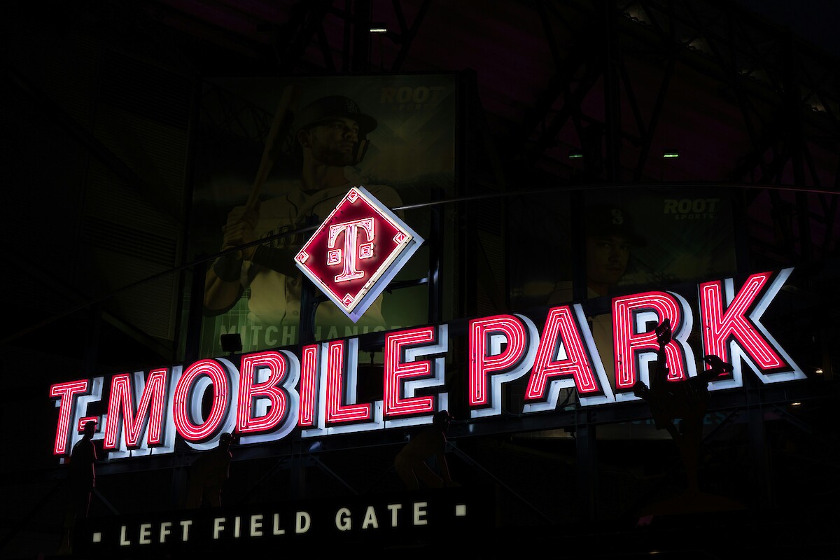 The image shows a T-Mobile Park sign with neon lights and a baseball player image in the background, at the Left Field Gate.