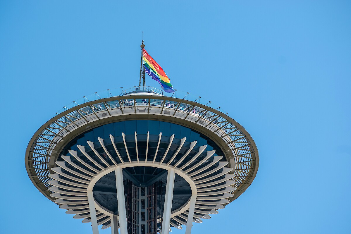 The image shows the top of the Space Needle with a rainbow flag flying on a clear day.