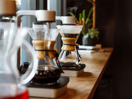 The image shows coffee being brewed using Chemex pour-over devices on a wooden counter.