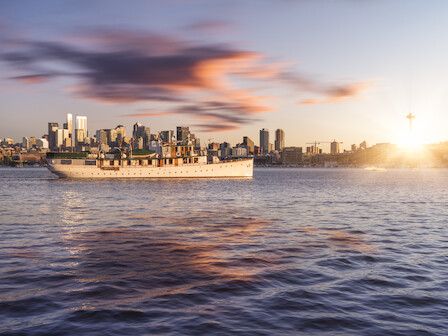 A ship sails in a calm bay with a city skyline and a sunset in the background, casting reflections on the water's surface.