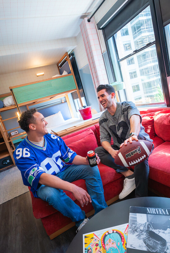 Two people in sports jerseys are sitting on a sofa; one holds a football, the other a drink. A table with magazines is in front.
