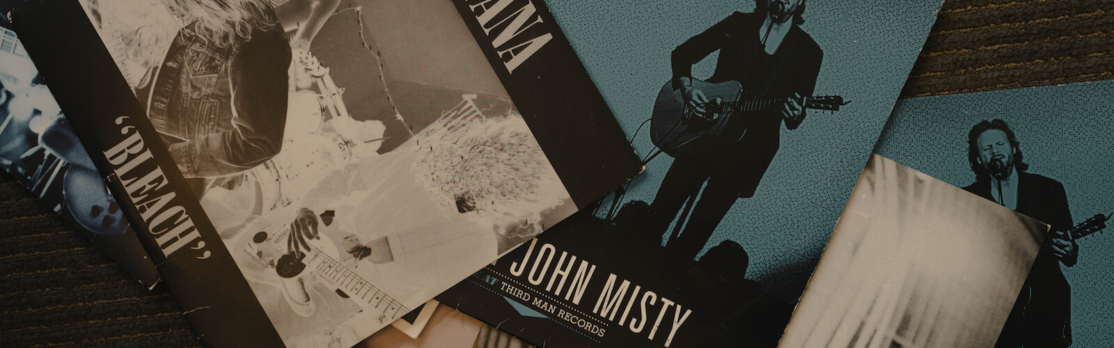 This image shows several vinyl record sleeves, including ones from Nirvana and Father John Misty, arranged on a dark surface.