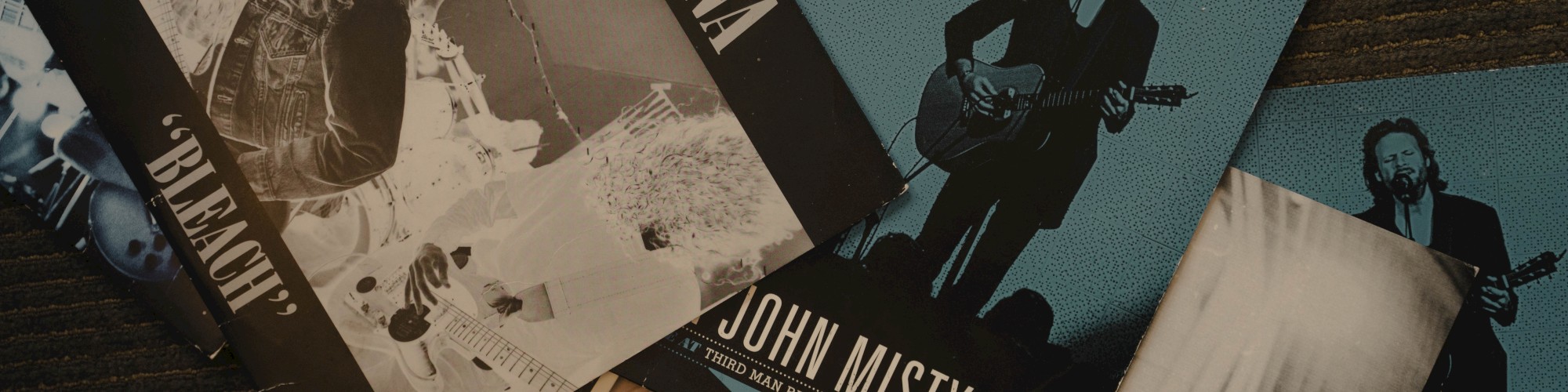 This image shows several vinyl record sleeves, including ones from Nirvana and Father John Misty, arranged on a dark surface.