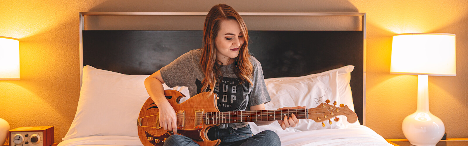 A person is sitting on a bed playing an electric guitar, with two bedside lamps turned on and some items, possibly books or records, nearby.