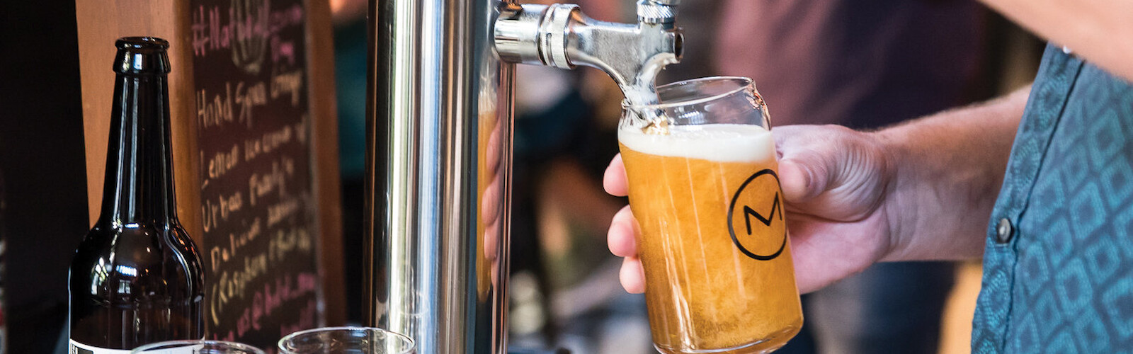 A person pours beer from a tap into a glass at a bar, next to other empty glasses and a bottle.