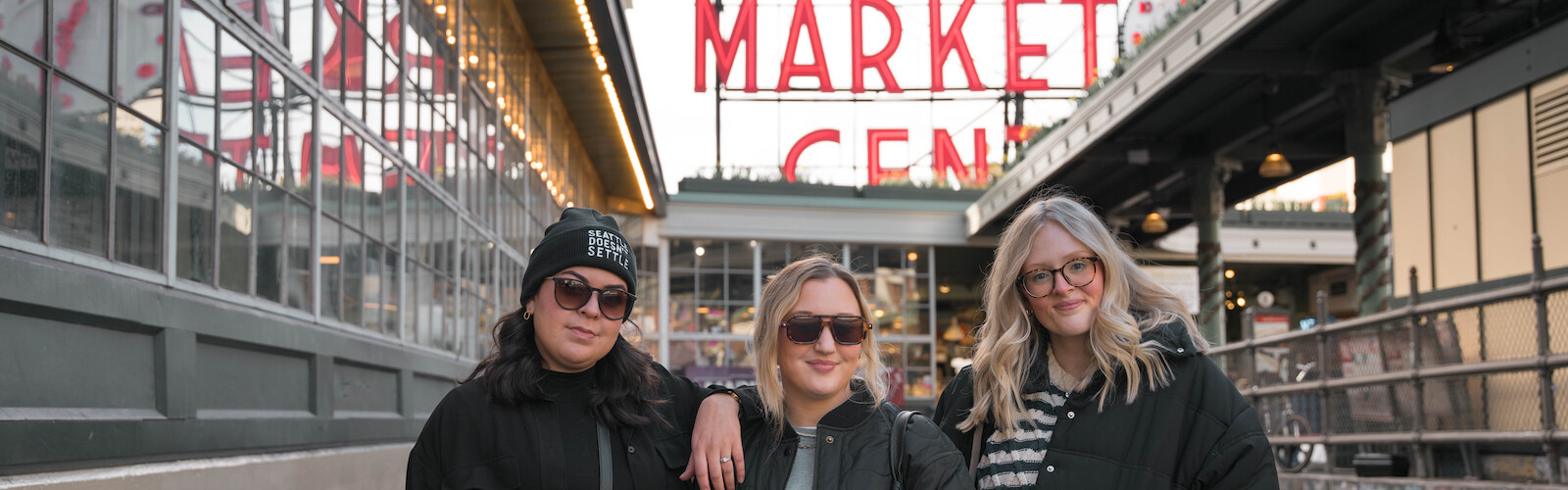 Three people stand in front of the Public Market Center sign, smiling and wearing sunglasses and jackets.