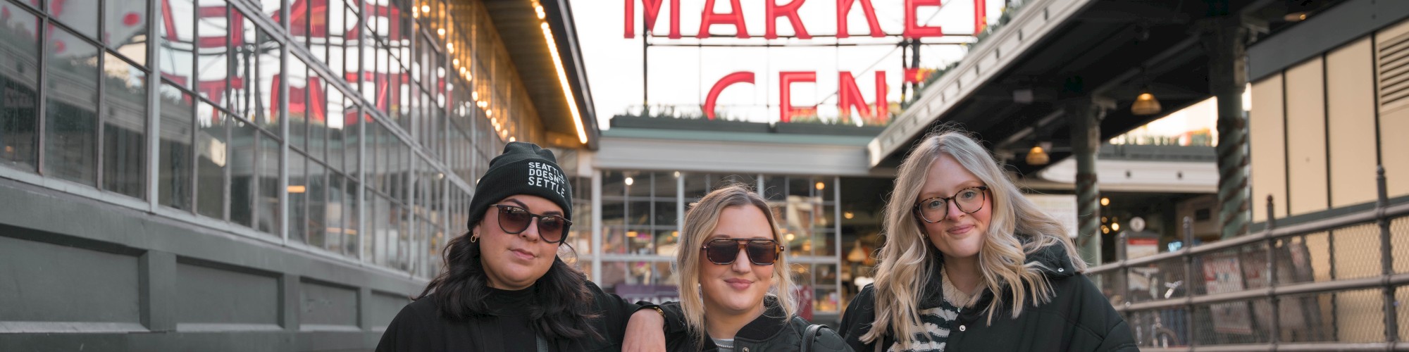Three people stand in front of the Public Market Center sign, smiling and wearing sunglasses and jackets.