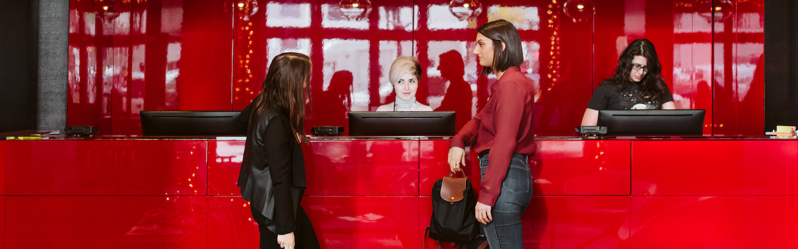Two people stand at a modern red reception desk, interacting with receptionists. One receptionist works on a computer.
