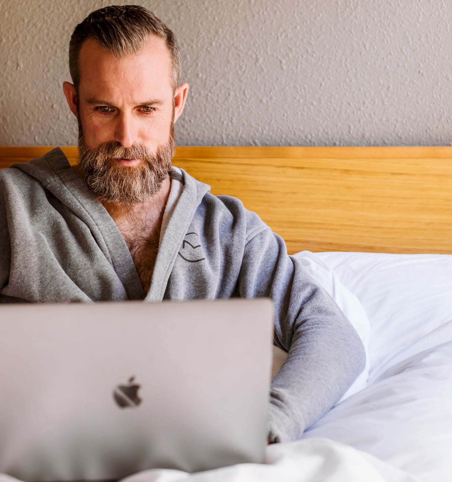 A bearded person is sitting in bed, wearing a gray robe, and using a silver laptop. The background shows a beige wall and a wooden bed headboard.