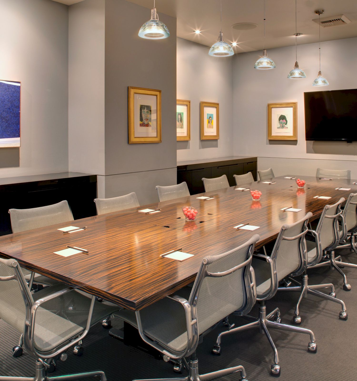 A modern conference room with a long wooden table, surrounding chairs, a wall-mounted TV, framed artworks, and pendant lights.