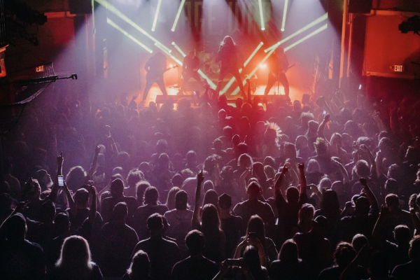 The image shows a crowded concert with a band performing on stage under vibrant lights, with the audience raising their hands, immersed in the music.