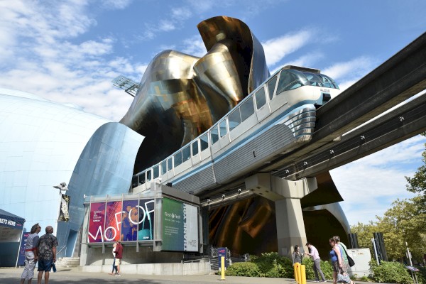 The image shows a modern building with a monorail passing through it, and several people walking nearby. The structure features a colorful facade.
