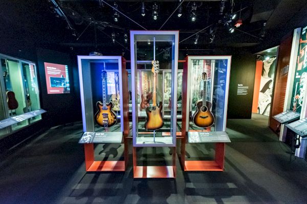 The image shows an exhibit of guitars displayed in glass cases within a museum setting. Numerous plaques and informational boards are visible.