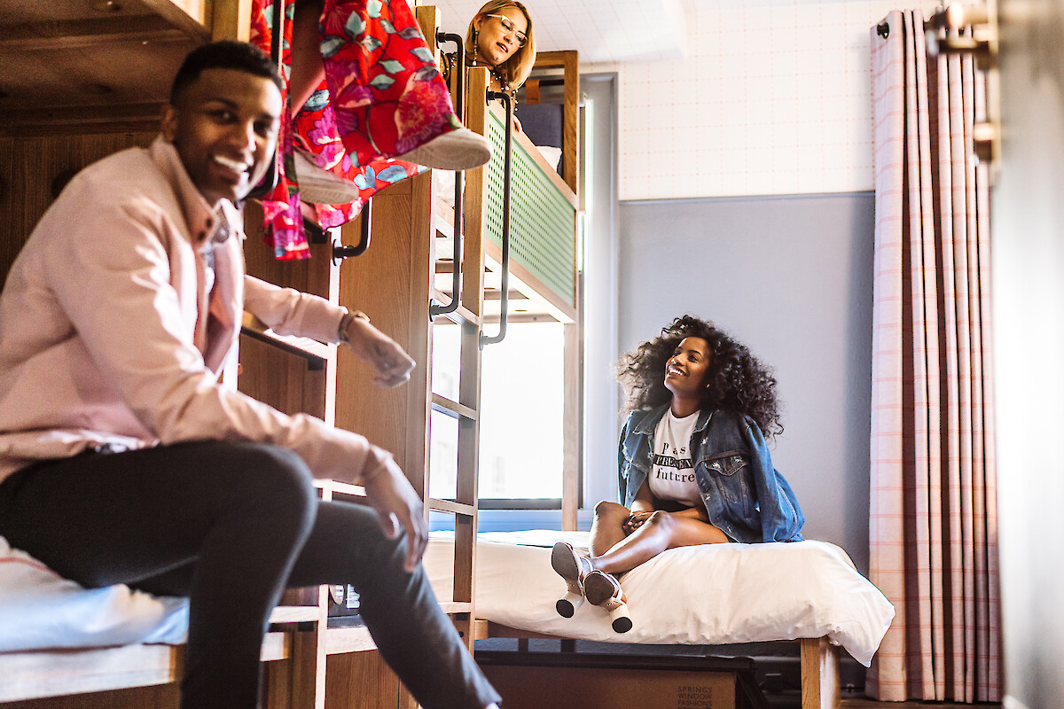 A group of friends enjoying time together in a room with bunk beds, one sitting on a bed, another sitting on upper bunk.