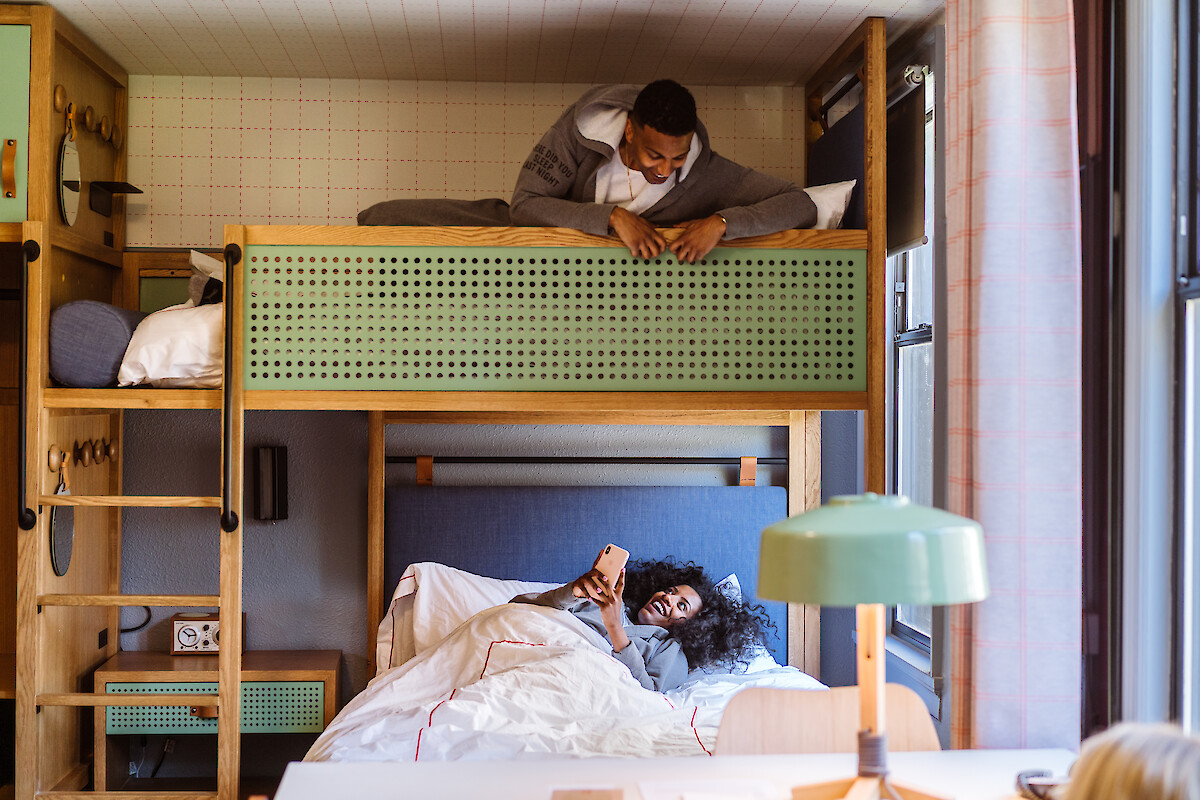 A person lies on a top bunk looking down, while another lounges on the bottom bunk holding a phone, in a cozy room with stylish decor.