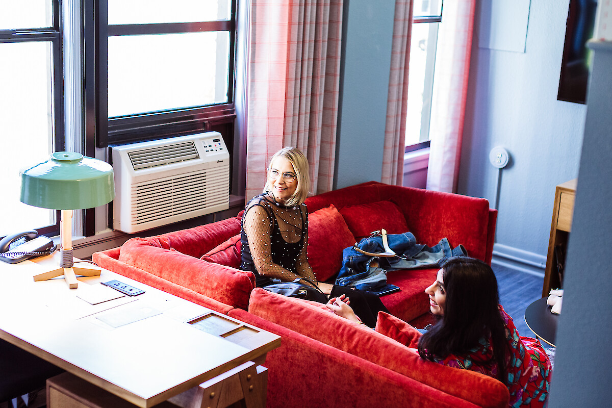 Two women sit on a bright red sofa in a cozy office living room, with a desk, lamp, and window AC unit nearby.