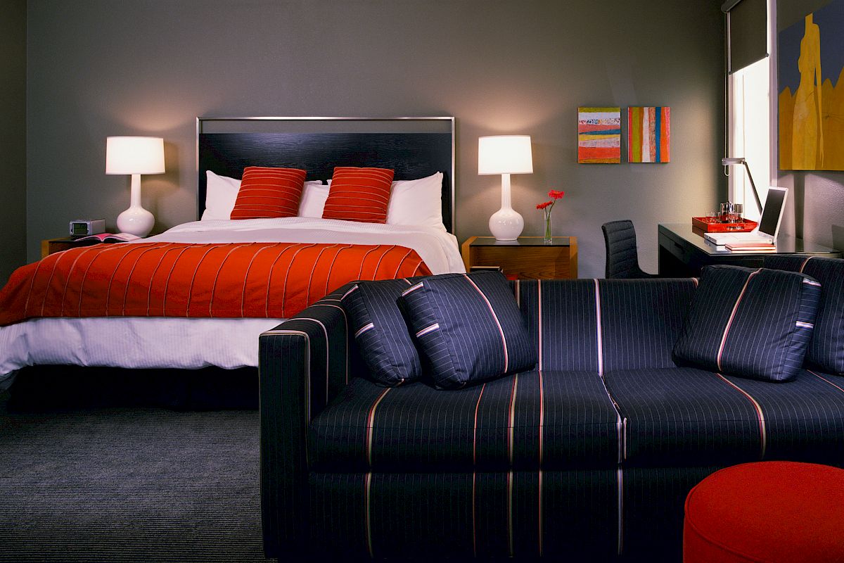 This image shows a modern bedroom with a double bed, two bedside lamps, a sofa, a desk, and colorful decor accents on the walls.