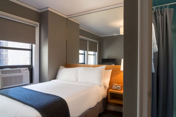 A cozy hotel room with a neatly made bed, nightstand, and wall-mounted air conditioner, featuring large windows and modern decor.