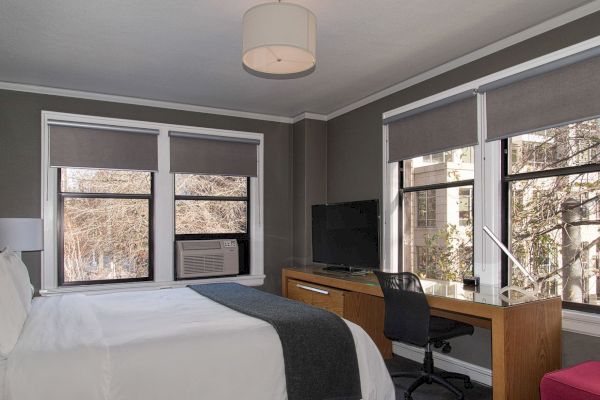 This image shows a modern bedroom with a bed, a desk with a TV and chair, and large windows with gray blinds. The room is well-lit.
