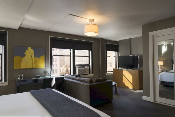 A modern hotel room features a bed, sofa, desk with a lamp, a TV, and artwork on the wall. It has large windows and a doorway to another room.