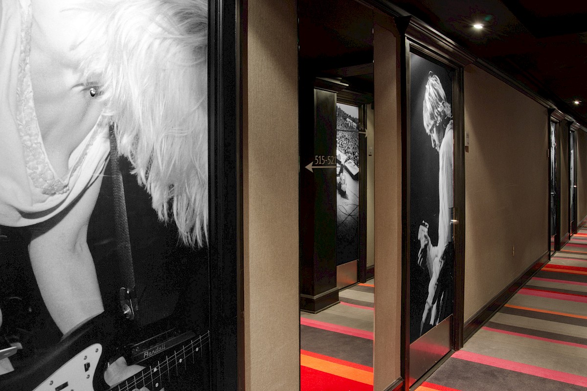 A hallway with large black-and-white photographs of people playing guitars on the walls, carpeted floor with red and gray stripes.