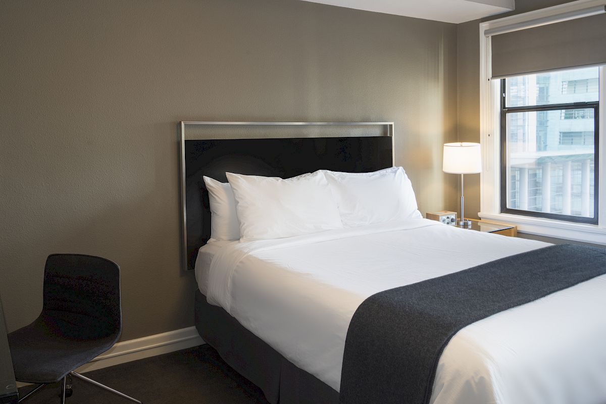 A neatly made bed with white linens and a dark blanket in a hotel room, featuring a black chair, a window, and a bedside lamp.