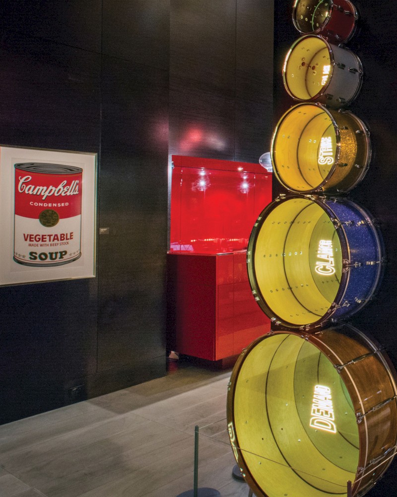 The image shows a vertical stack of colorful drums, a Campbell's soup poster, and a red illuminated area in the background.