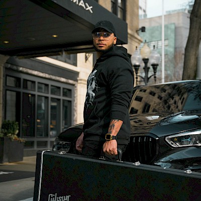 A person stands outside Hotel Max holding a Gibson guitar case, dressed in dark clothing with a black cap, next to a black vehicle.