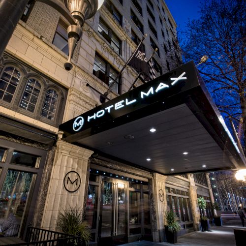 The image shows the entrance of Hotel Max, featuring its illuminated sign and canopy, with a mix of modern and historic architectural elements.