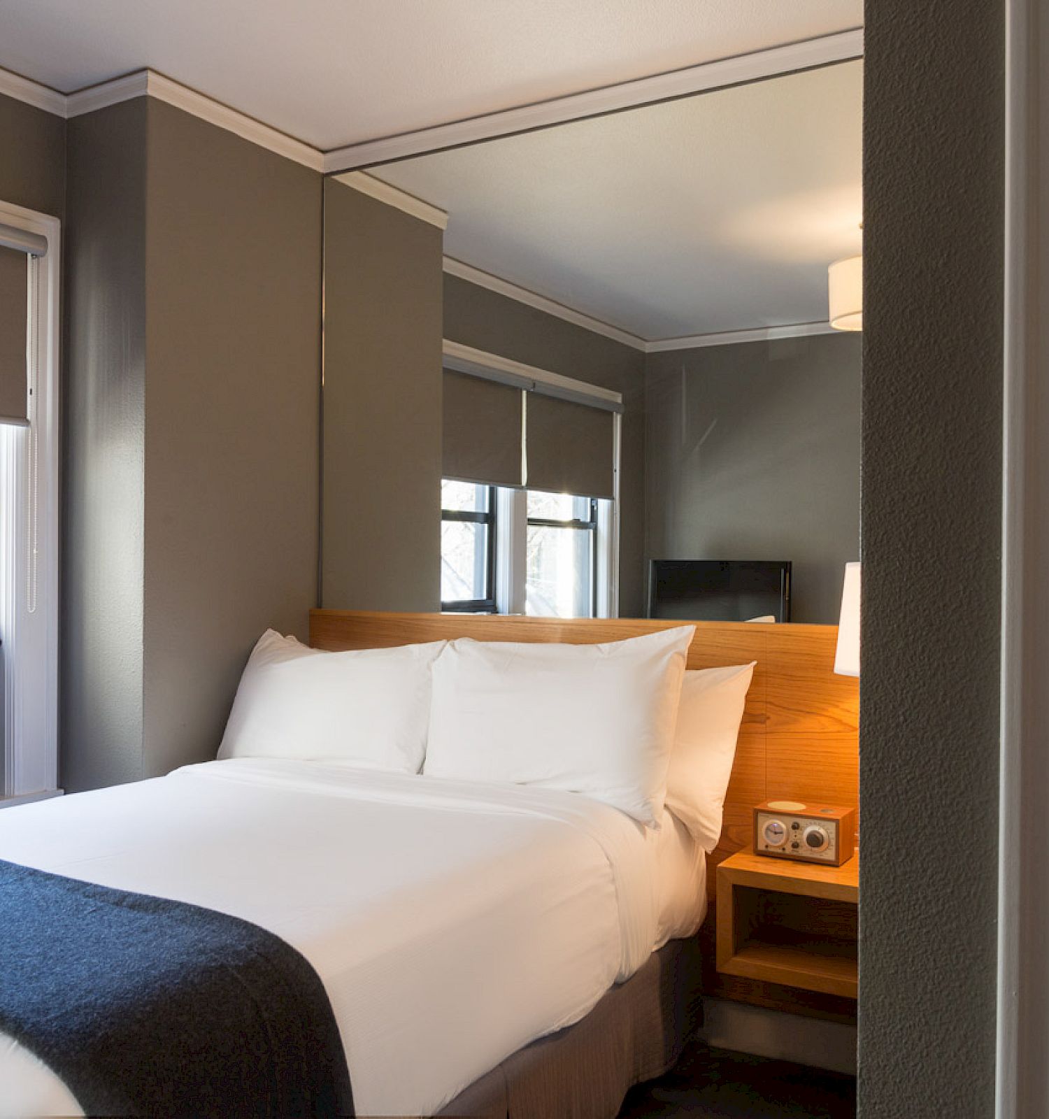 The image shows a small, modern hotel room with a neatly made bed, a nightstand, a window, air conditioning, and a mirror.