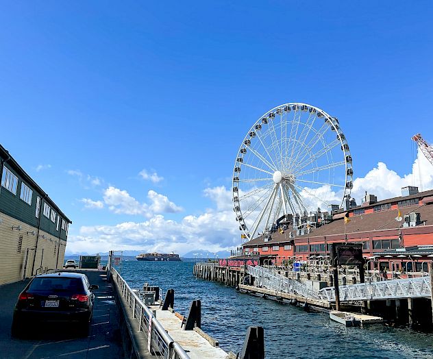 A waterfront scene with a Ferris wheel, docked boats, and a car on the left side, under a clear blue sky.