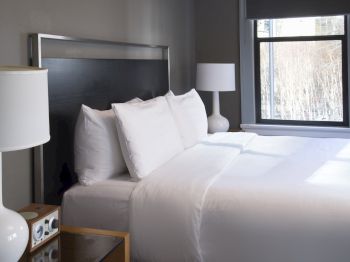A neatly made bed with white linens, two bedside tables with white lamps, and a window letting in natural light are in the room.
