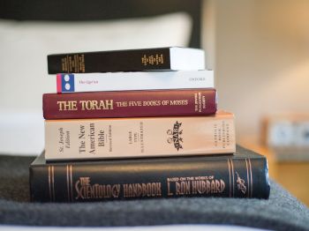 The image shows a stack of five books, including The Qur'an, The Torah, The New American Bible, the Bhagavad Gita, and The Scientology Handbook.