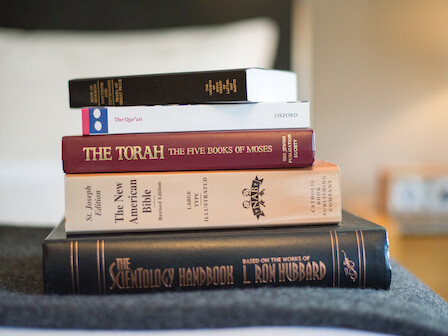 The image shows a stack of five books, including The Qur'an, The Torah, The New American Bible, the Bhagavad Gita, and The Scientology Handbook.