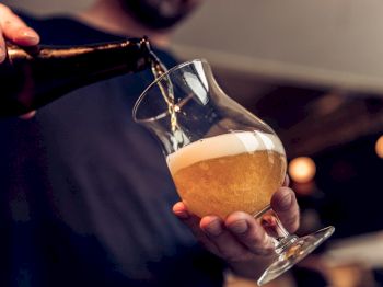A person is pouring beer from a bottle into a tulip glass, which is partially filled with frothy beer.