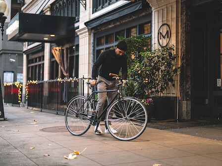 A person is standing on a sidewalk, adjusting or inspecting a bicycle in an urban setting with decorated storefronts in the background.