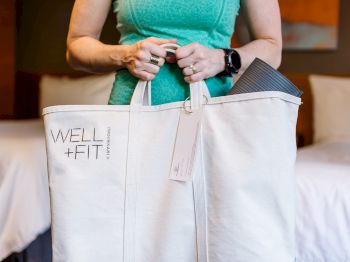 A person in a green top holds a white Well + Fit bag with a yoga mat visible inside, standing in a room with a bed in the background.