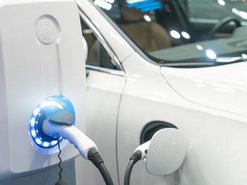 The image shows an electric vehicle being charged at a charging station, with the charger plugged into the car and a glowing light indicating charging.