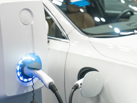 The image shows an electric vehicle being charged at a charging station, with the charger plugged into the car and a glowing light indicating charging.