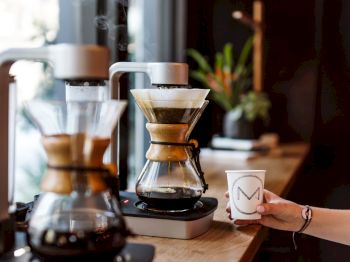 The image shows a person holding a coffee cup next to a coffee-making apparatus, with a plant in the background.
