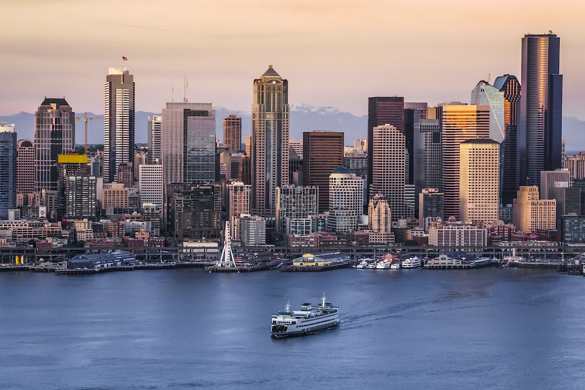 A city skyline at sunset with various high-rise buildings and a ferryboat traversing the water in the foreground, set against a mountainous backdrop.