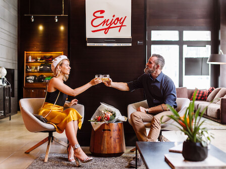 Two people toast with drinks in a stylish living room; a sign says “Enjoy” in red above them as they sit and chat.